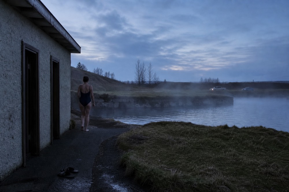 A visitor in a swimsuit walking from a changing room doorway toward steaming geothermal water at dusk