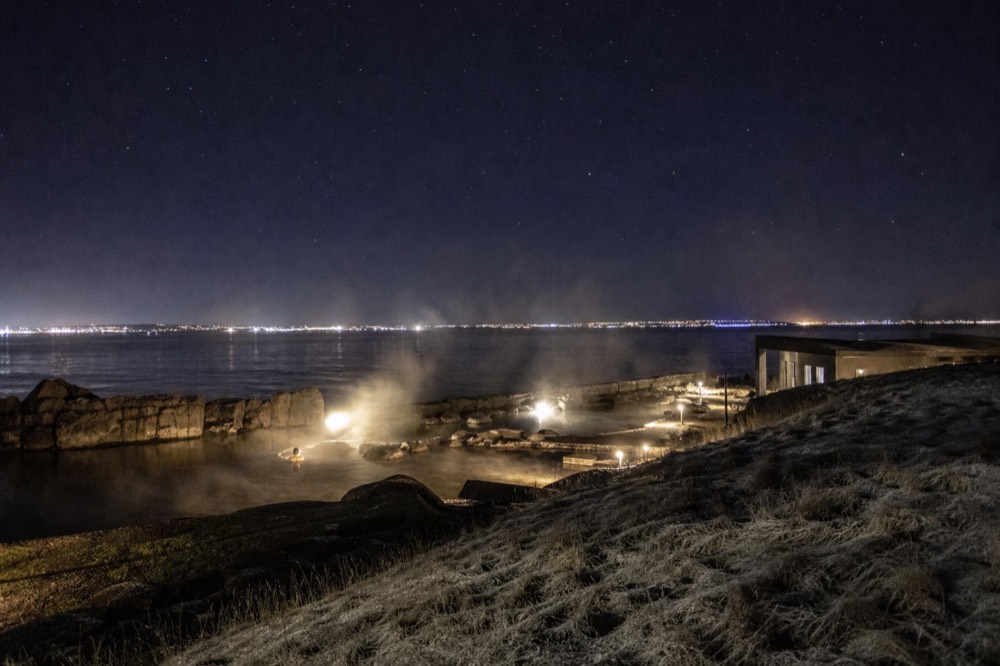 A dark night sky above a steaming geothermal pool at the edge of a town