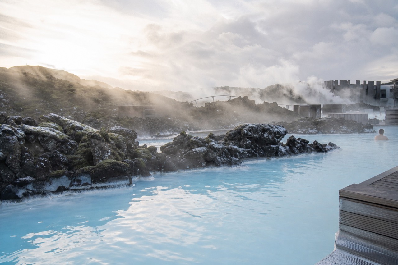 Steam rising from pale blue geothermal water beside a dark lava ridge