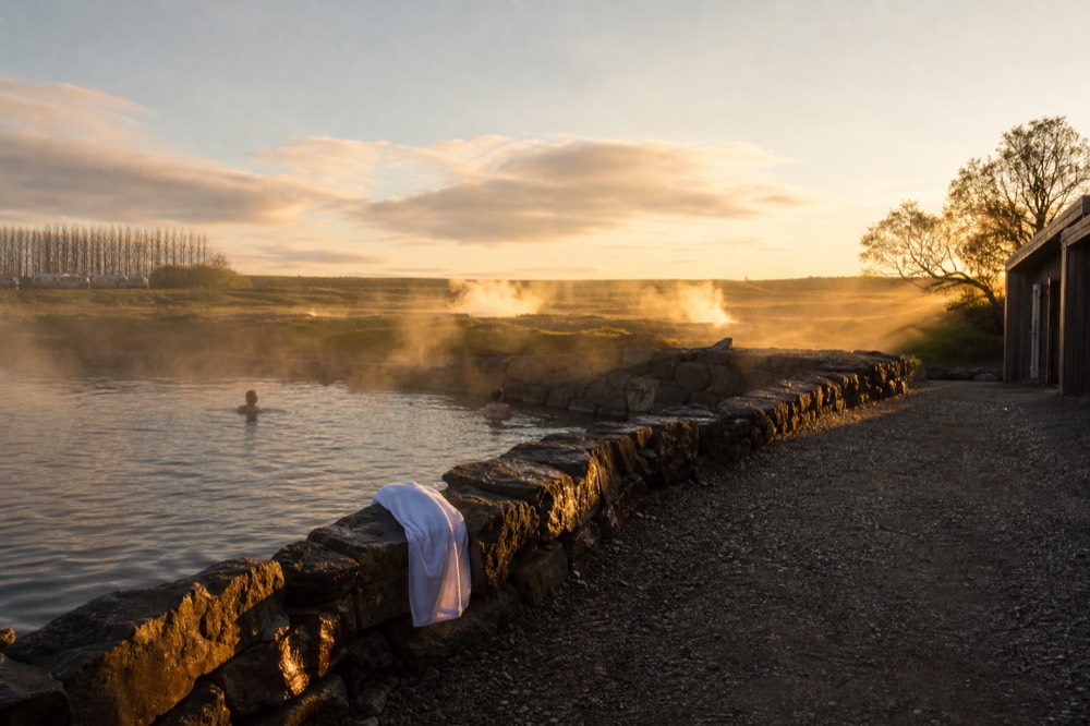 A historic geothermal pool at golden hour with steam catching low light over flat farmland