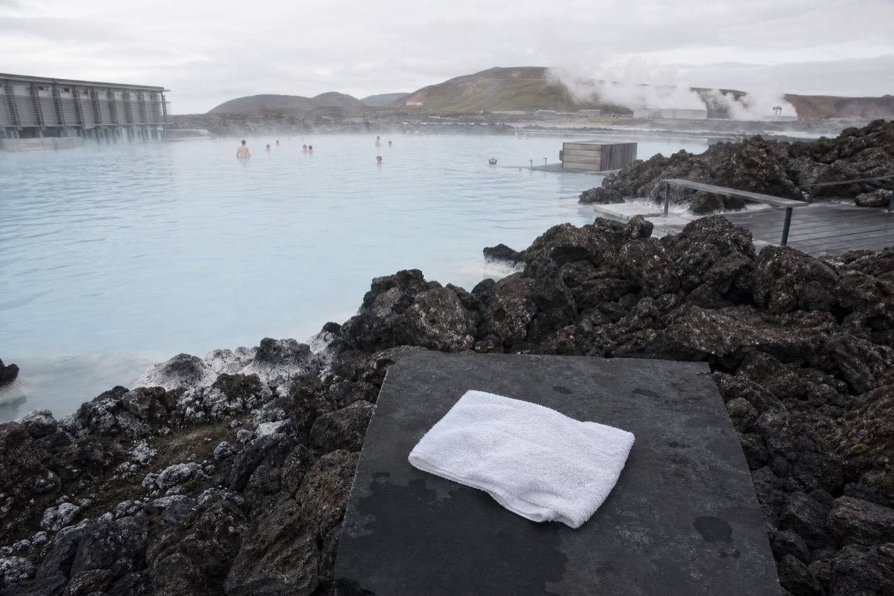 Steam rising off a pale blue geothermal lagoon surrounded by dark lava rock under a soft sky