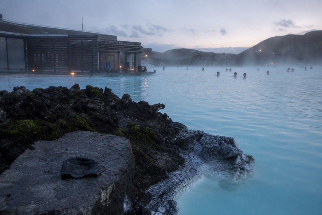 Steam rising from pale blue geothermal water in a lava field at dusk