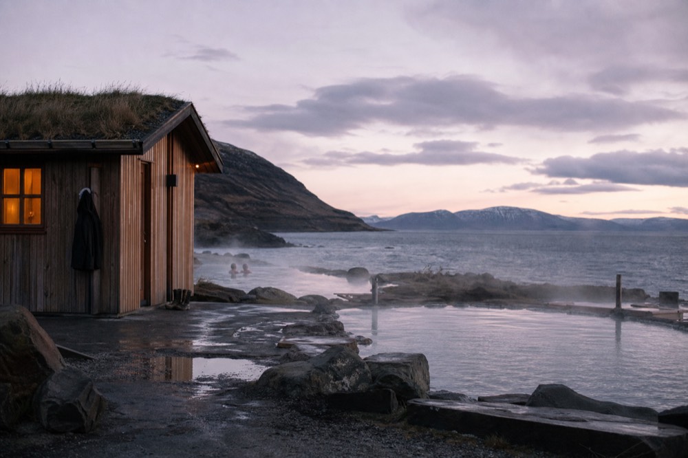 A wooden sauna building beside steaming pools on a fjord shoreline at dusk