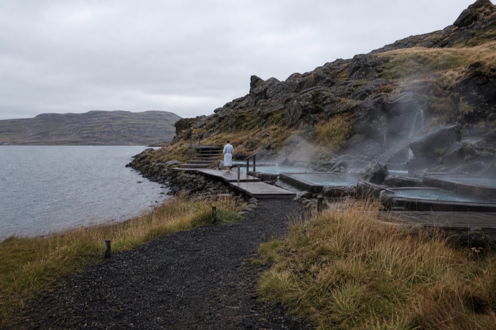 Steaming geothermal pools built into a dark hillside beside a calm fjord