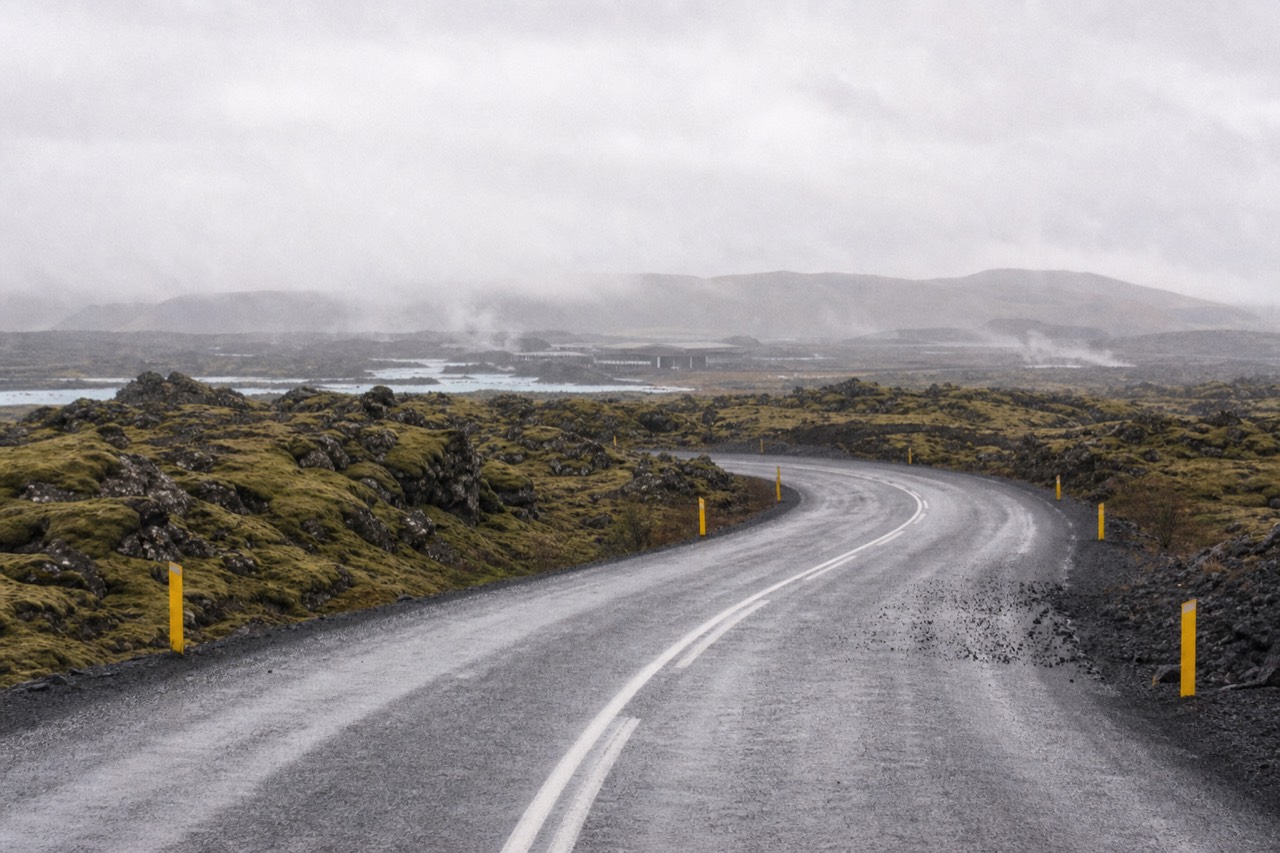 A empty two-lane road curving through a moss-covered lava field under a grey sky