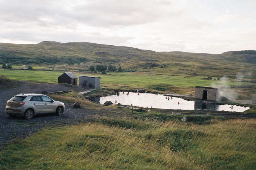 A geothermal pool beside a rural village with fields and low hills in the background