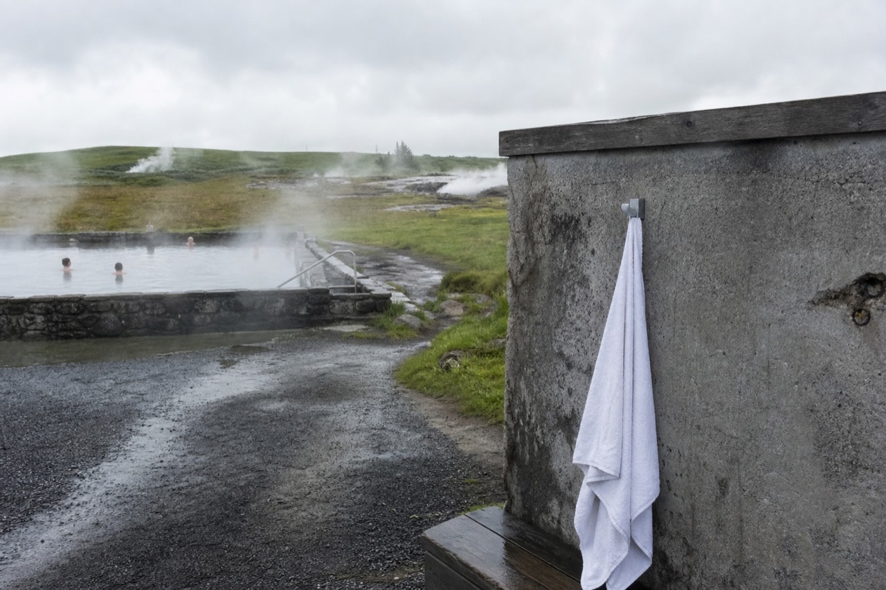 Steam rising from a geothermal pool surrounded by gravel paths and small hot springs on a misty day