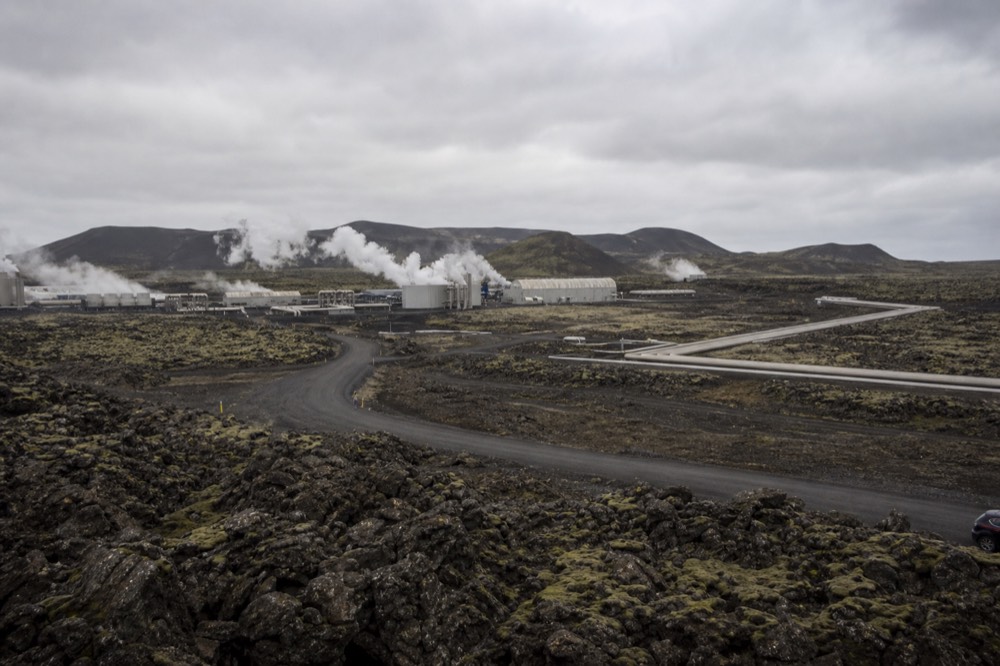 A steaming geothermal power station on a flat lava field under a wide grey sky