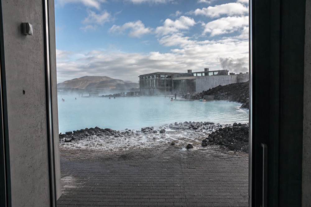 A pale blue geothermal lagoon with a modern building and steam drifting across still water