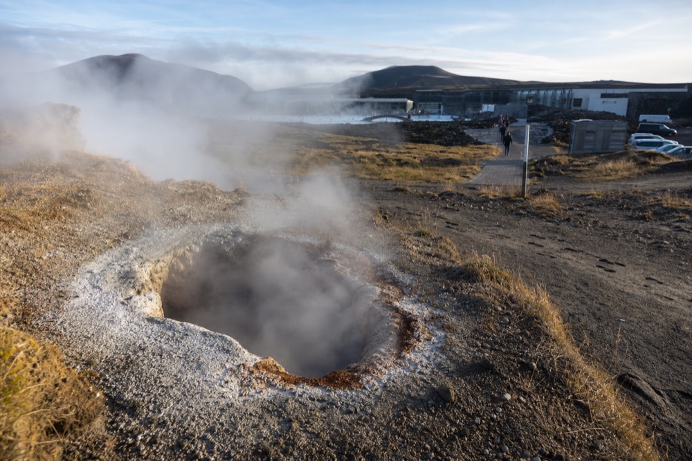 A steaming geothermal vent in a gravel field with mineral deposits around its edge