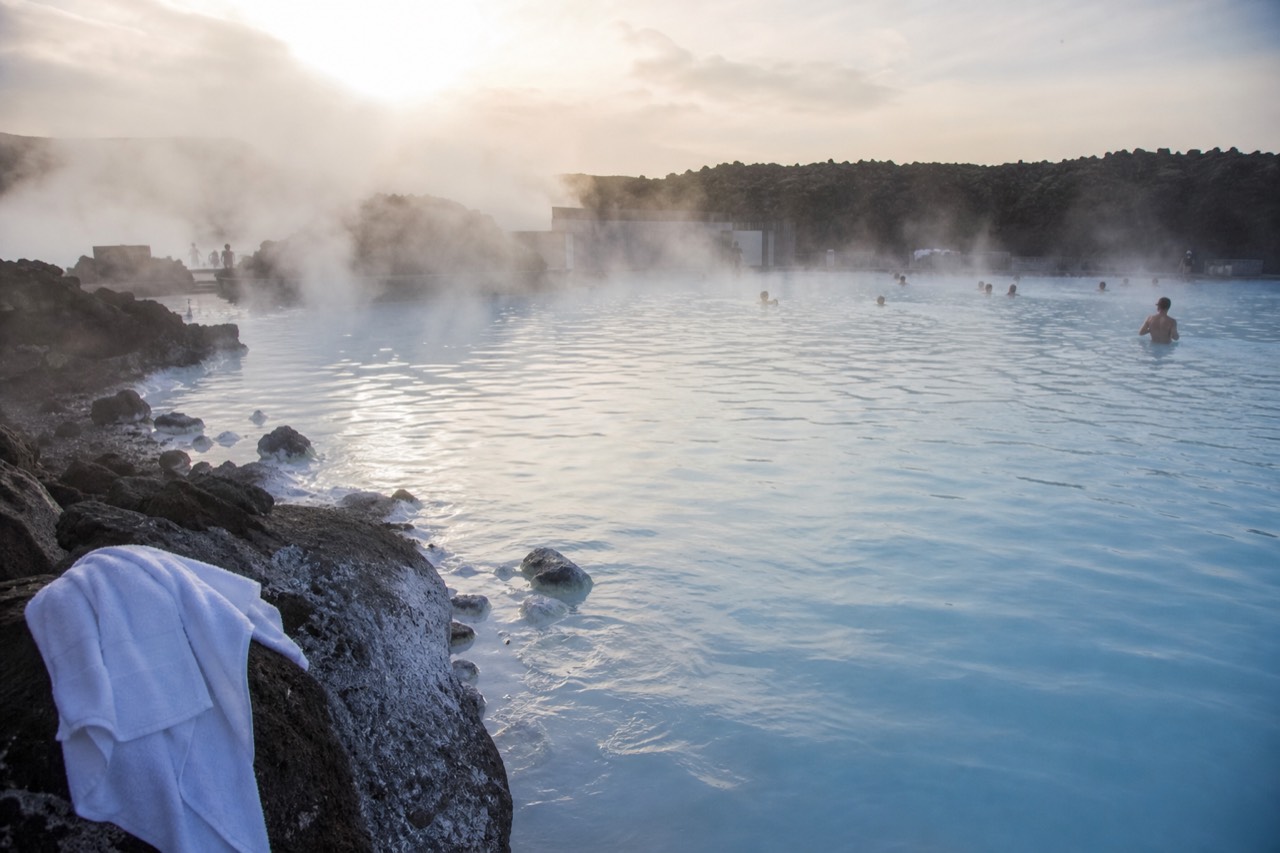Steam rising off the pale blue water of a geothermal lagoon surrounded by dark lava rock