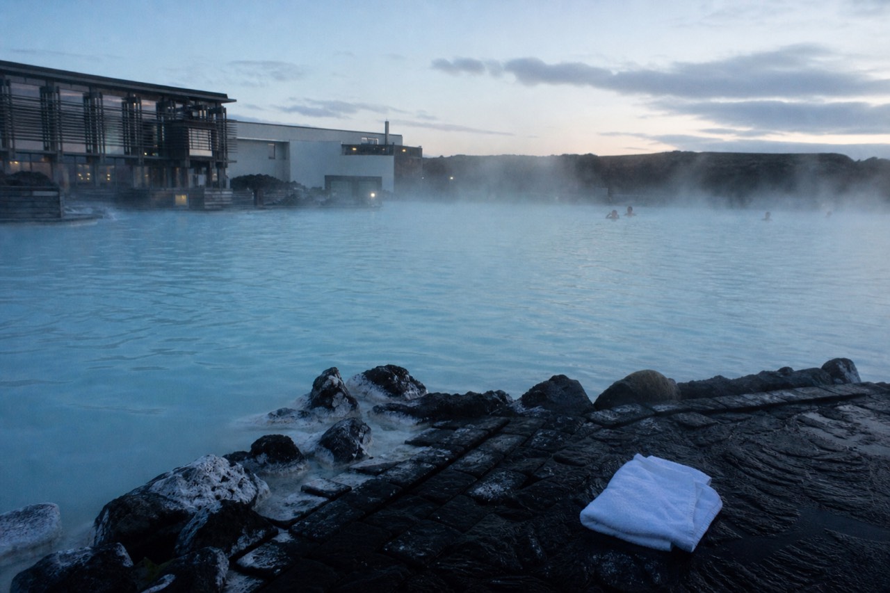 Steam rising from a geothermal lagoon beside dark volcanic rocks at dusk