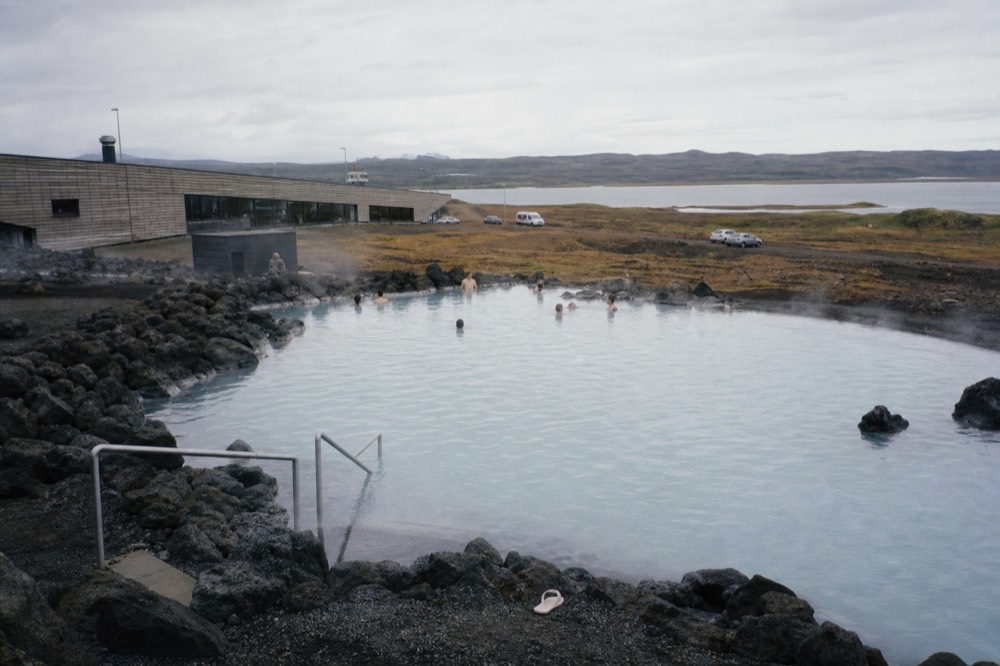 A steaming geothermal pool edged with dark lava rock under a pale sky
