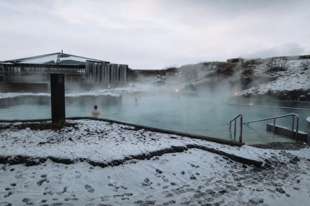 A steaming geothermal pool beside a snowy lava field under a pale winter sky