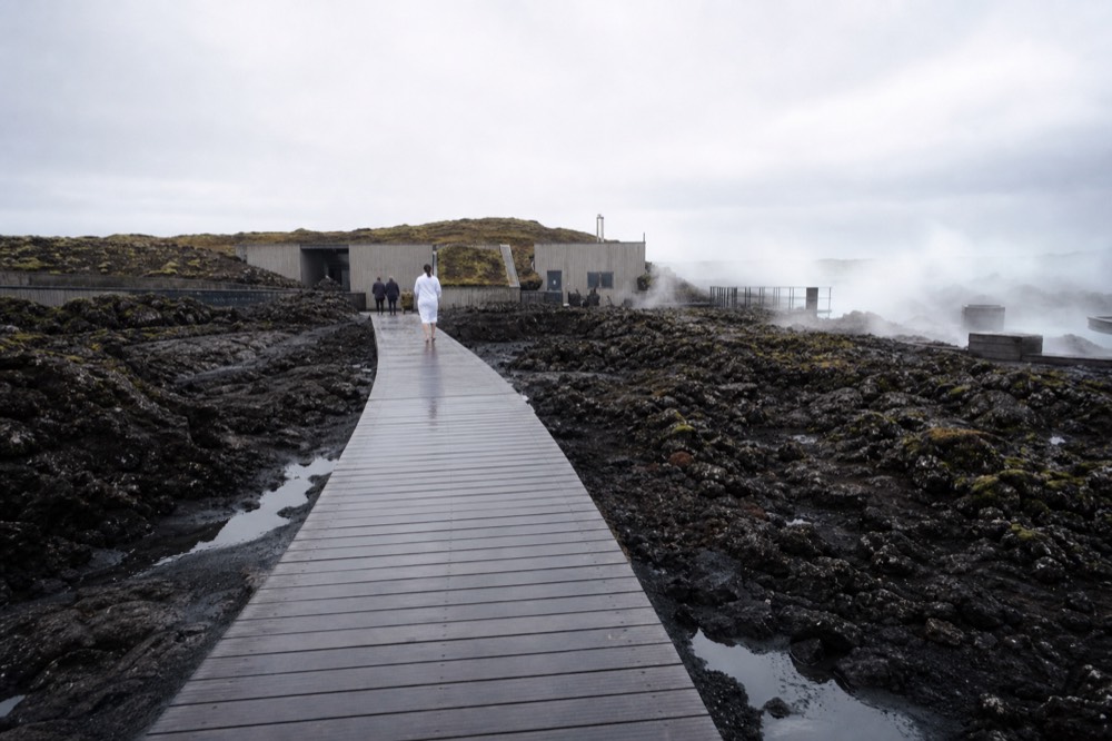 A wooden walkway leading across a lava field toward a low spa building