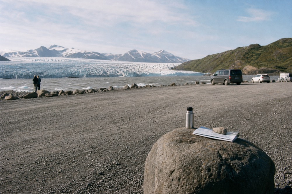 Empty gravel parking area beside a glacial lagoon with a low glacier in the distance