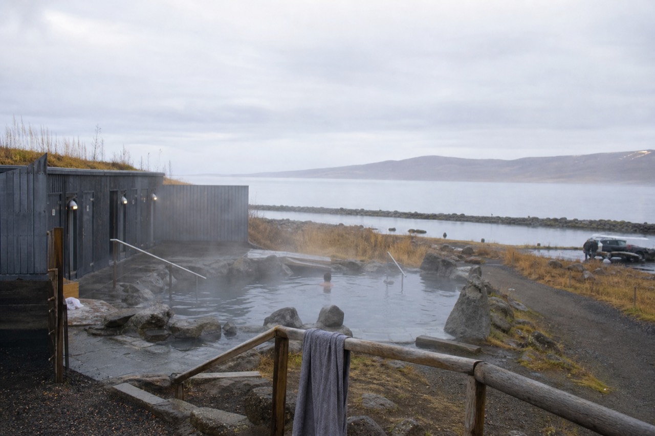 Steam rising from geothermal baths on the shore of a lake with mountains behind