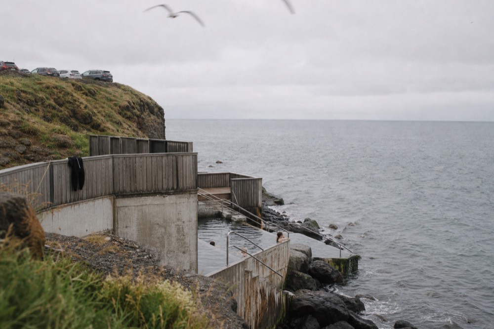 A modern timber bathing complex on a coastal cliff with steam rising from water against an open sea horizon