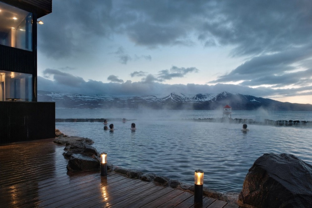 A calm infinity pool at dusk with steam drifting across the surface toward distant mountains