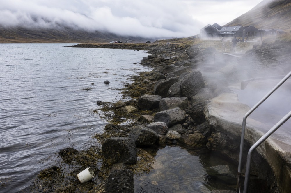 Steam rising from geothermal pools on a dark shoreline beside a calm fjord