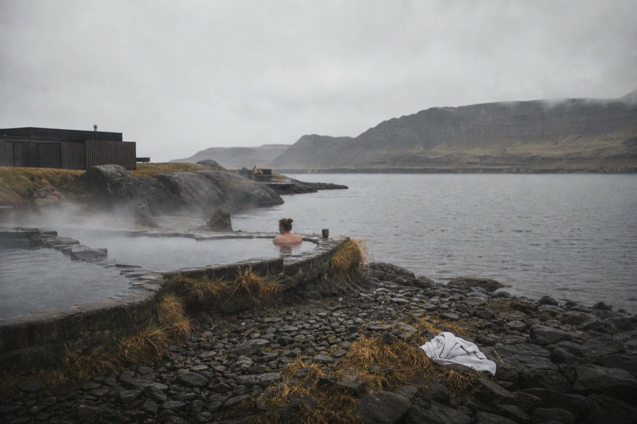 Steaming geothermal pools on a dark shoreline with mountains across a fjord