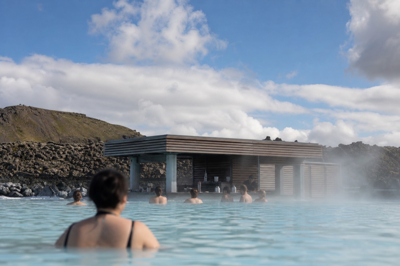 A steaming geothermal lagoon with a swim-up bar and guests relaxing in the water, surrounded by a rugged lava landscape under a bright sky.