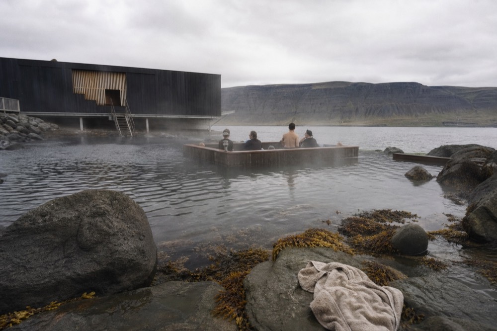 A wooden swim-up bar with stools partly submerged in a warm outdoor pool overlooking a fjord