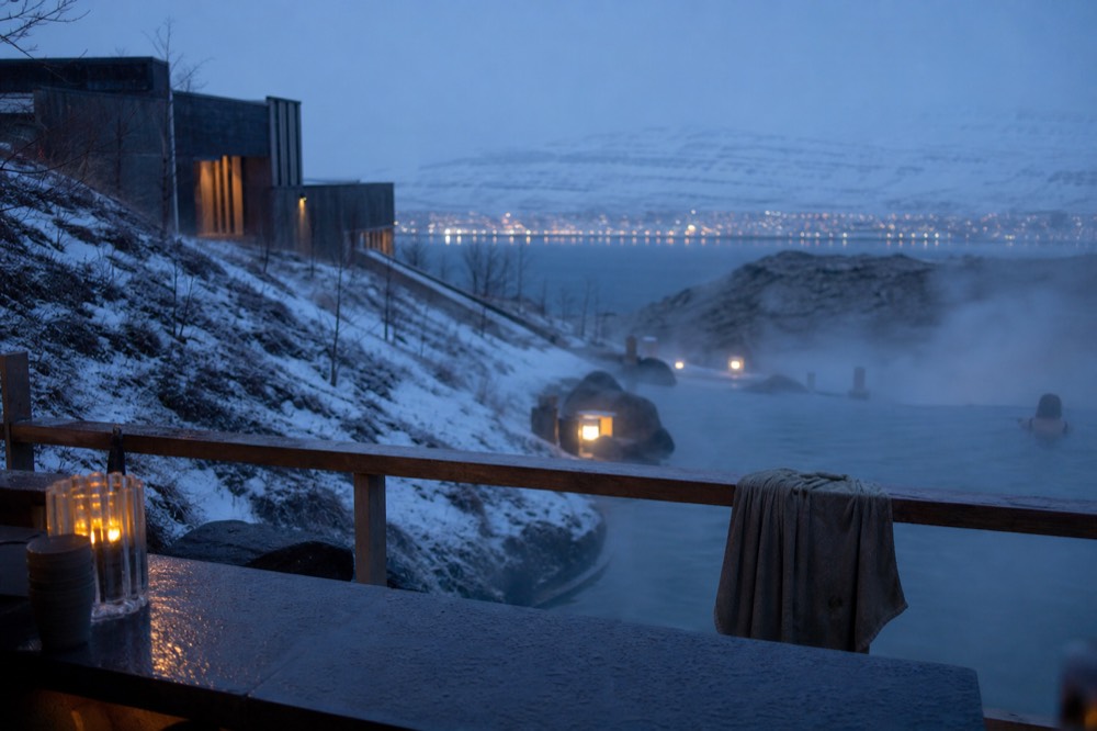 An evening view across a fjord from a warm outdoor pool with a bar counter in the foreground