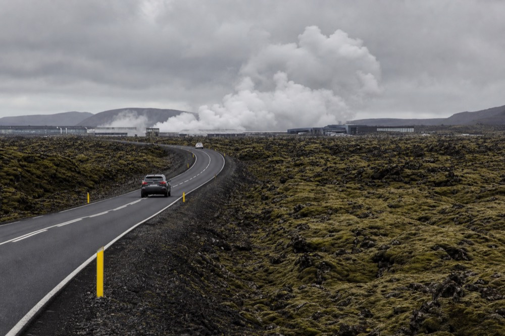 A road winding through moss covered lava fields toward a distant geothermal steam plume