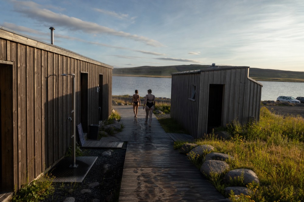 Travellers in swimsuits walking between wooden buildings at a geothermal spa in evening light