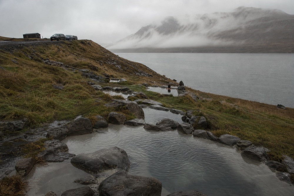 Steaming natural pools terraced down a grassy slope toward a calm grey fjord