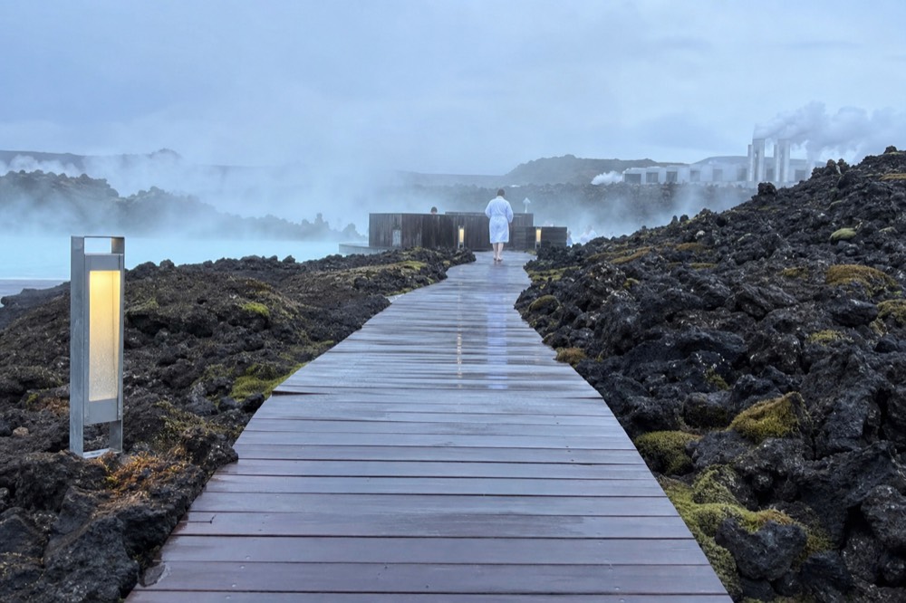 A wooden walkway crossing dark volcanic rock toward a steaming lagoon