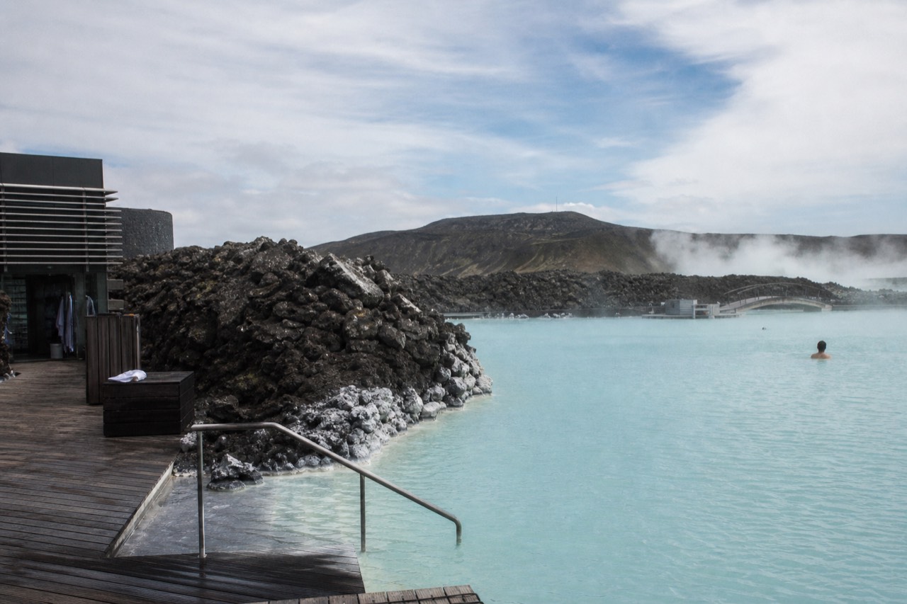 Steam rising over a pale blue geothermal lagoon surrounded by dark lava rock under a wide sky
