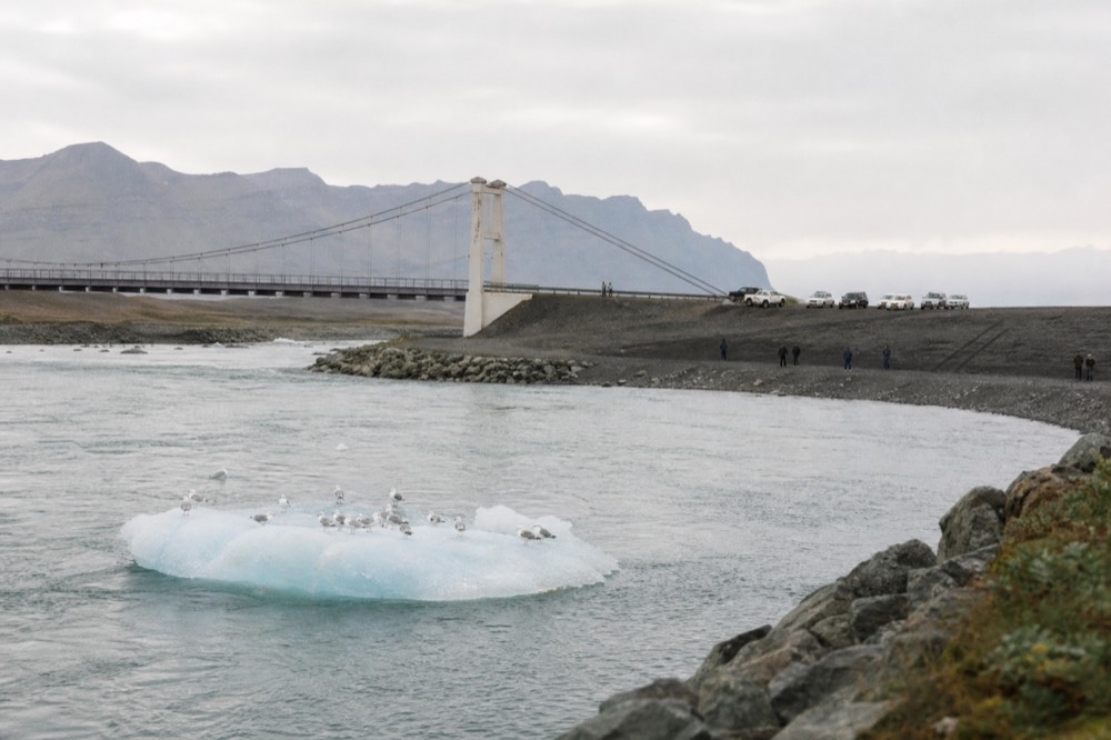 A wide view of a glacier lagoon with a bridge and black sand shore in the distance