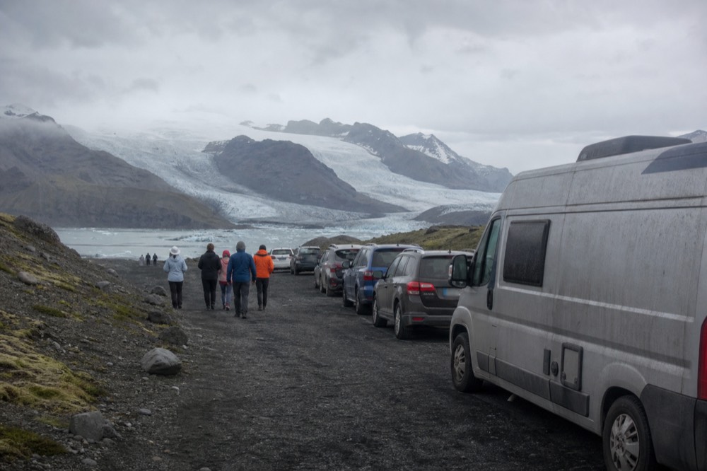 A gravel parking area beside a lagoon with people walking toward the water