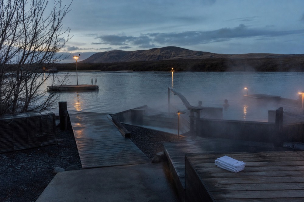 Steam rising from geothermal pools built out over a calm lake at dusk