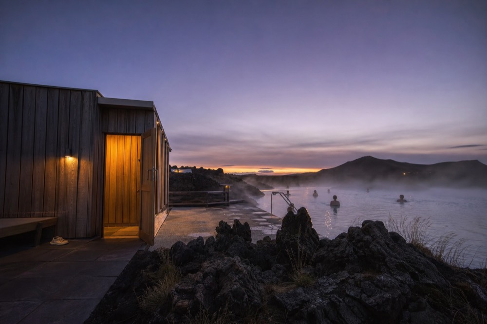 A modern timber bathing building with a steaming outdoor pool at dusk