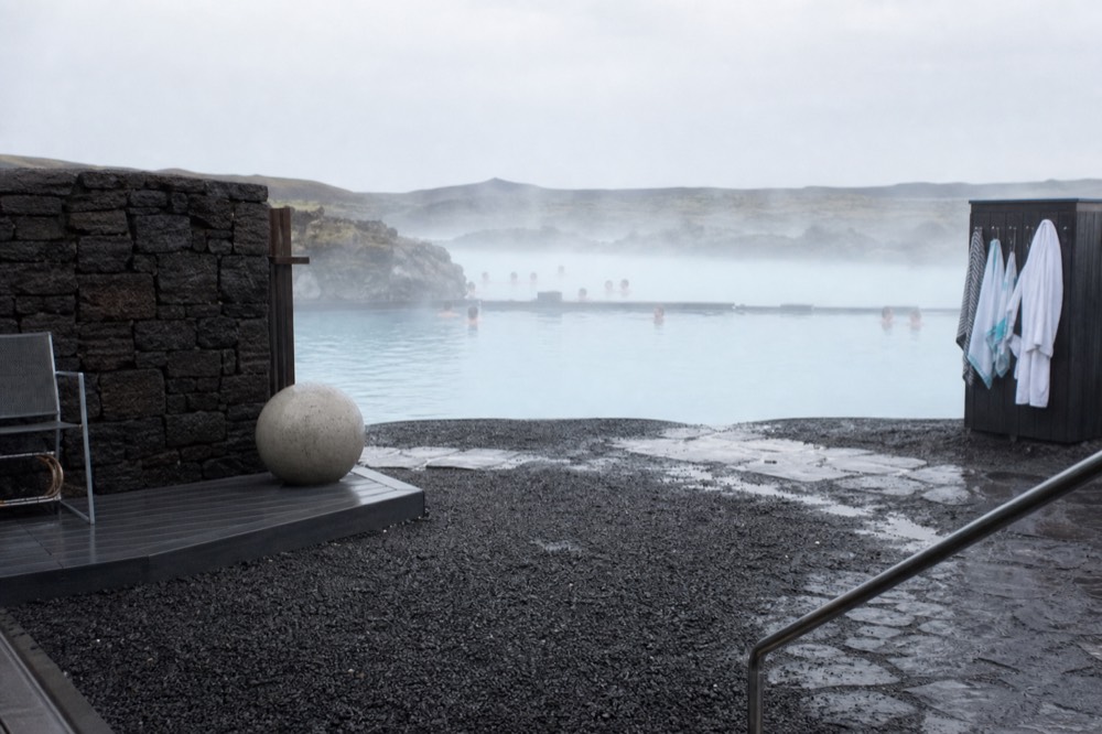 A quiet poolside terrace with steam drifting past dark rock walls under a pale sky