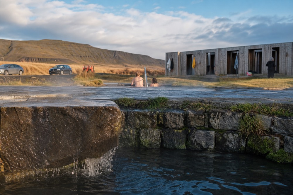 A steaming geothermal pool edged with dark stone at the base of a green valley