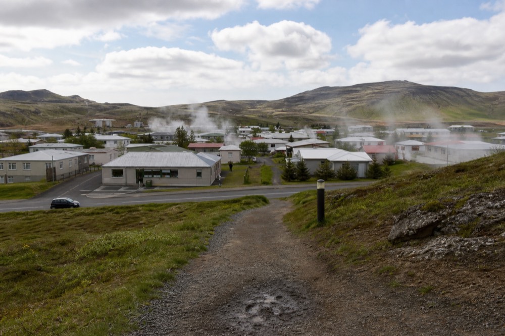 A small Icelandic town with green hills and drifting steam behind it