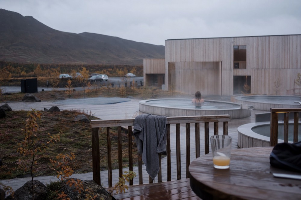 A wooden deck and steaming pools beside a modern bathhouse building