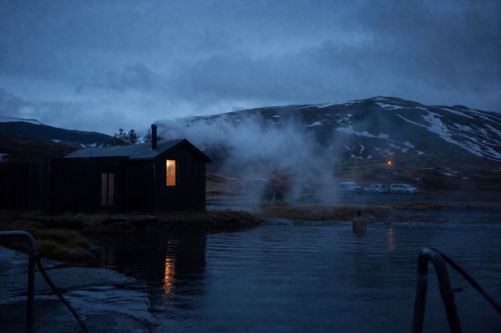 Steam rising from a sauna building beside a still geothermal pool at dusk