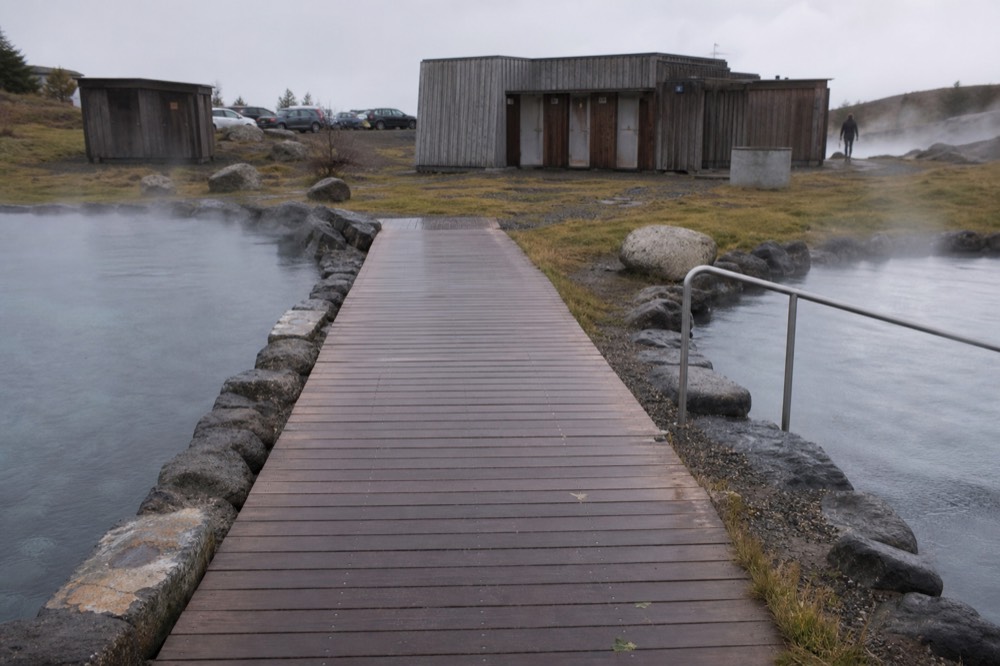 An empty wooden walkway between steaming pools under an overcast sky