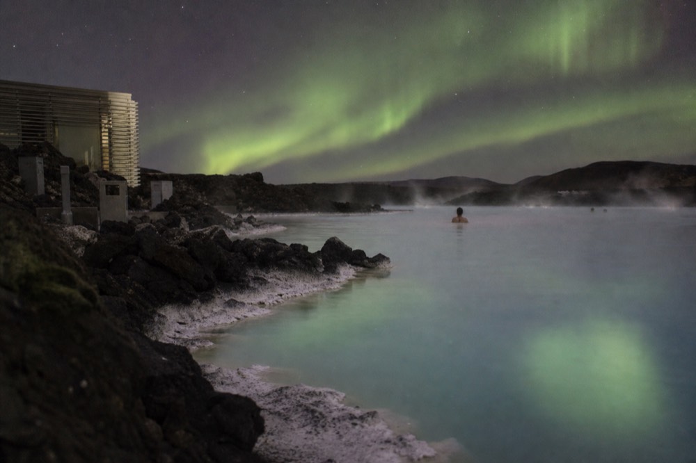 A calm geothermal pool at night reflecting a green aurora over dark hills