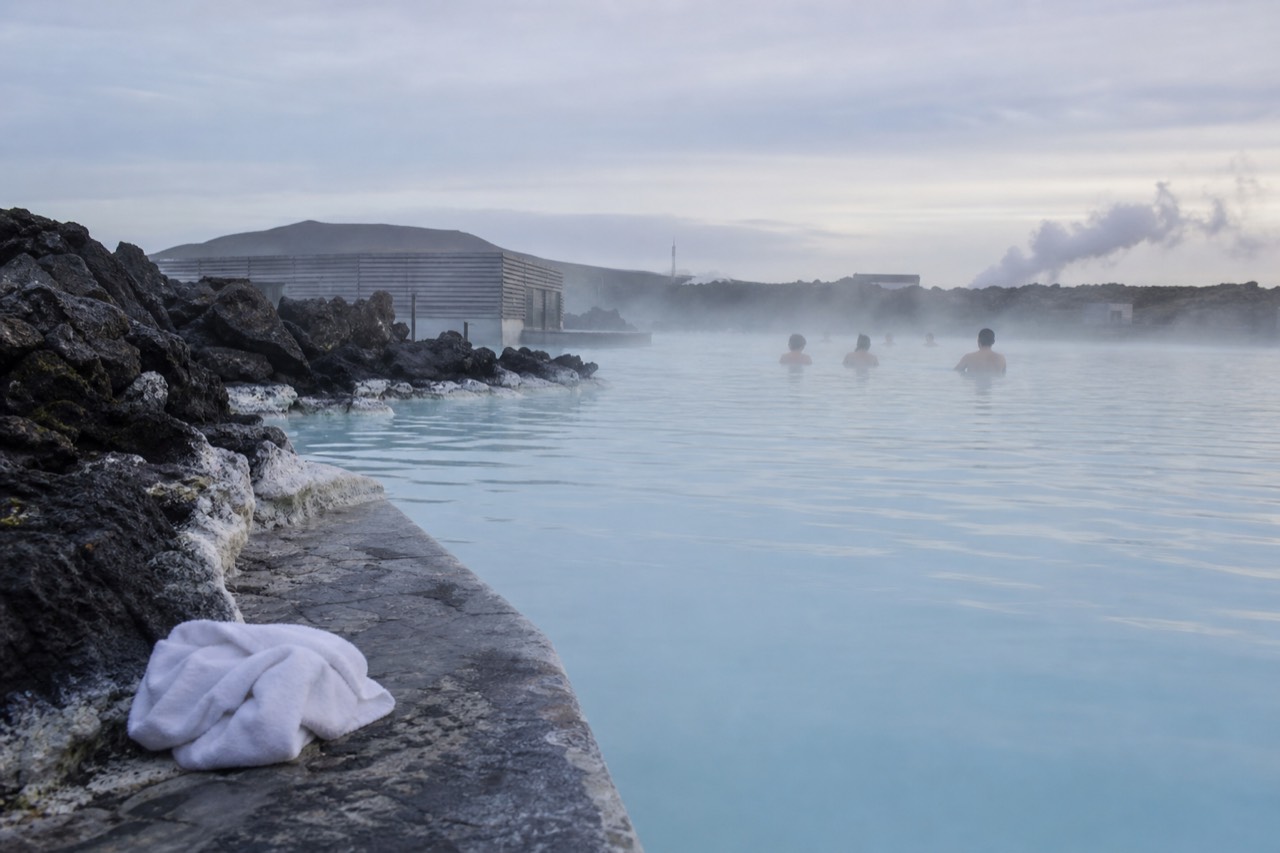 A steaming geothermal lagoon in Iceland at dusk, pale blue water edged by dark volcanic rock, with bathers visible in the mist and low hills beyond.